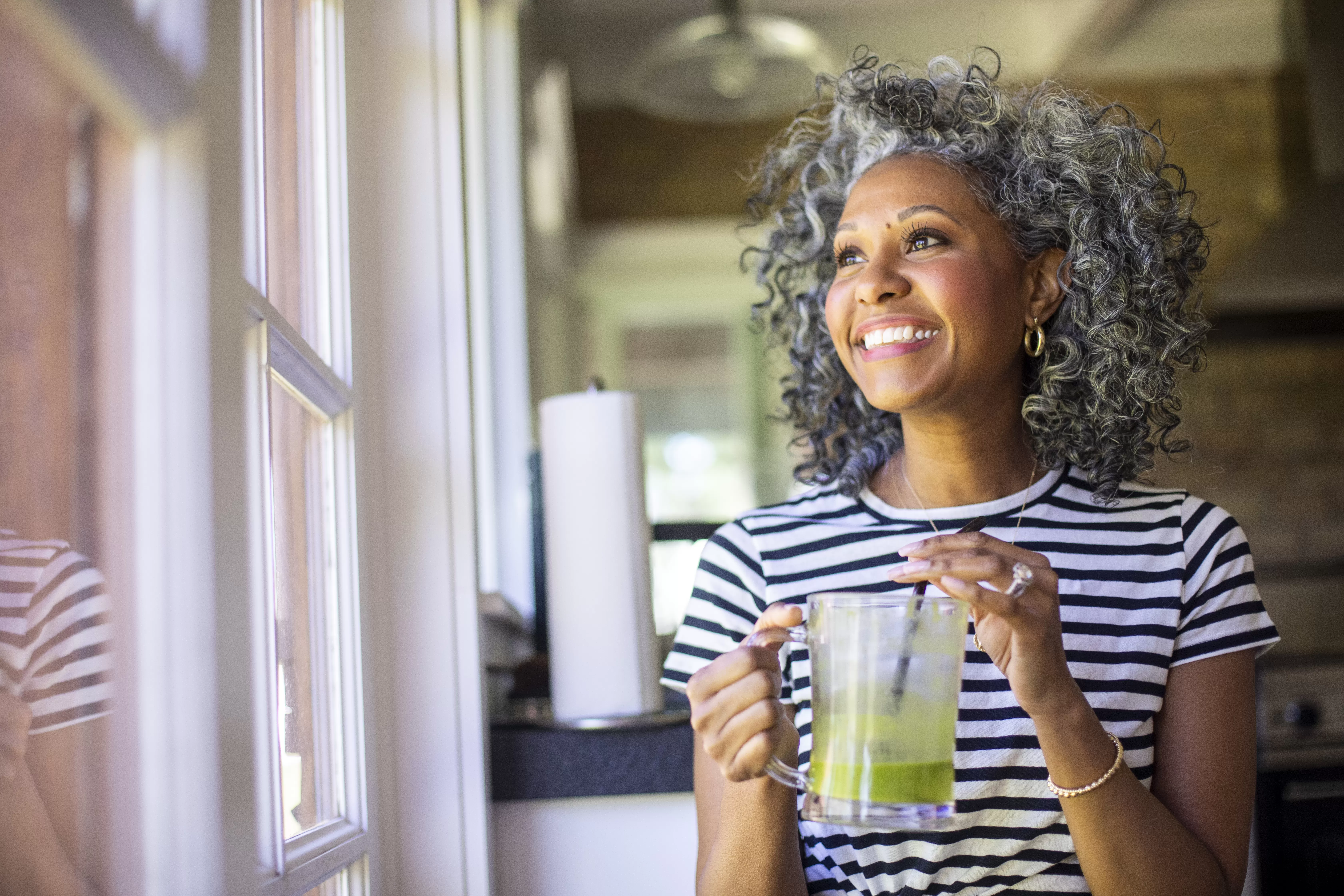 Mulher negra sorrindo de cabelos grisalhos, blusa listrada branco e preto, copo com líquido verde, copo transparente.